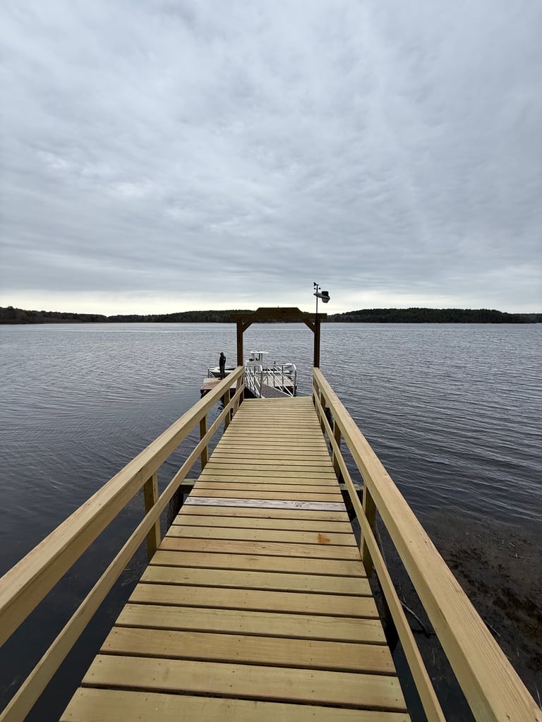 Wooden dock extending into calm gray water under an overcast sky with a covered boathouse structure at the end