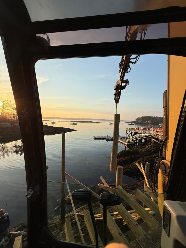View from inside a boat or ship crane at sunset, looking out over calm harbor waters with sailboats and shoreline.