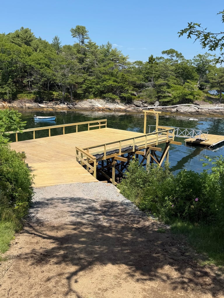 Wooden dock and boat launch extending into calm river water surrounded by green trees and rocky shoreline on a clear day