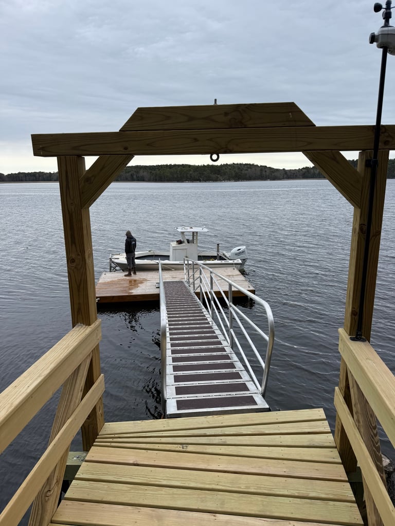Wooden dock with pergola overlooking a boat ramp and white pontoon boat on calm lake water