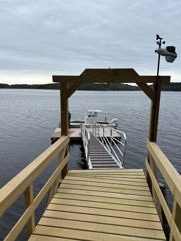 Wooden dock with pergola extending into calm lake water, with a boat docked below and weather station visible on the roof