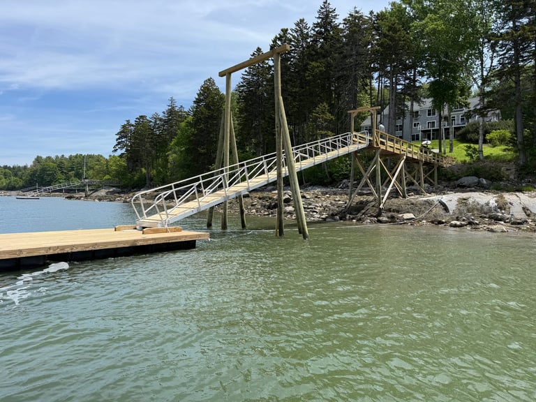 Wooden pedestrian bridge with rope railings crossing calm turquoise water, connecting a dock to a forested shoreline with residential buildings visible in the background