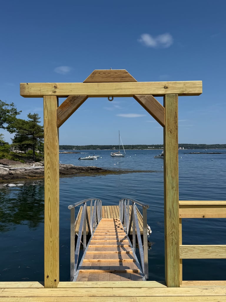 Wooden pergola frames a long dock leading to calm blue water with sailboats in the distance on a clear day