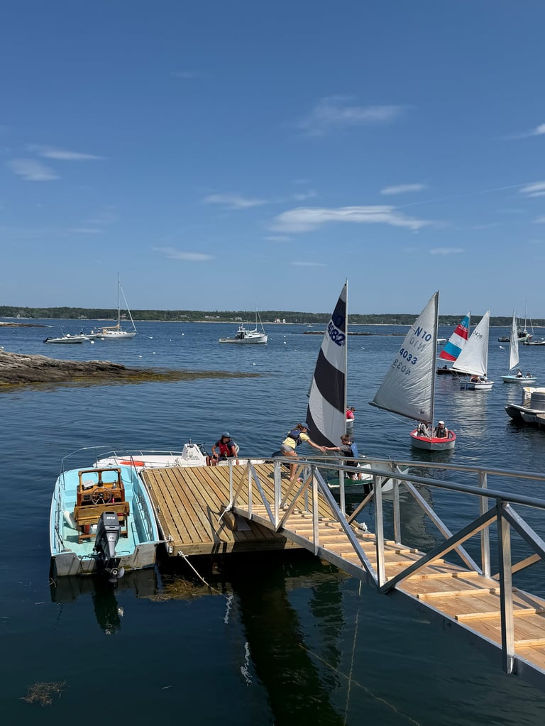 Sailboats moored in a blue harbor with people on a wooden dock in the foreground under a clear sky