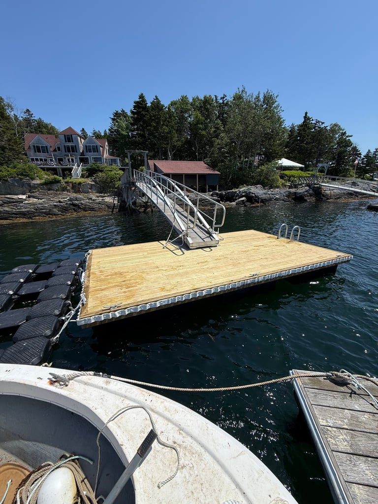 Wooden dock platform on blue water with boat, footbridge, and residential cottage visible on rocky shore among trees