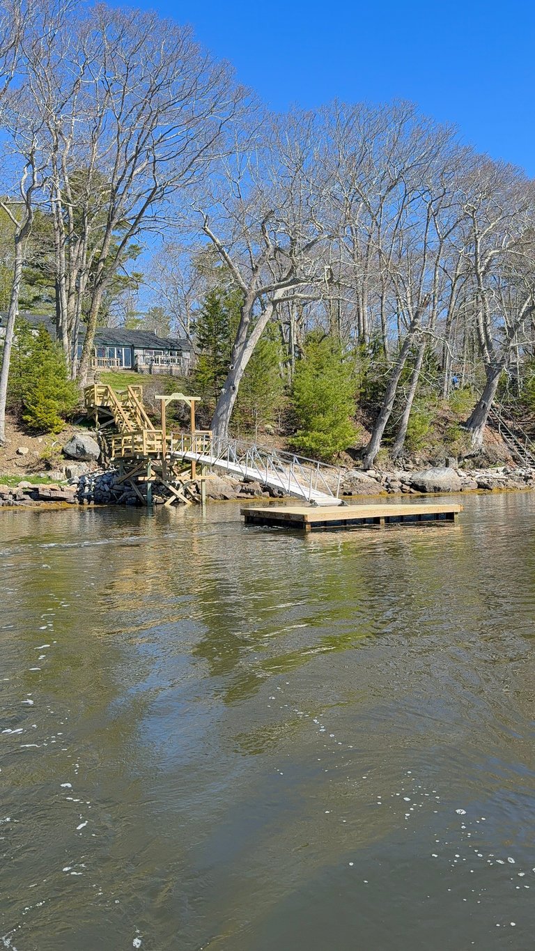 Waterfront house with wooden dock and boat launch on river surrounded by bare trees and green conifers on sunny day