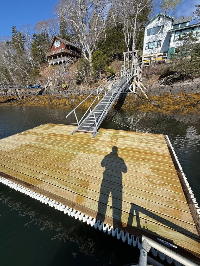 Person standing on wooden dock casting shadow, with waterfront houses and metal walkway bridge in background