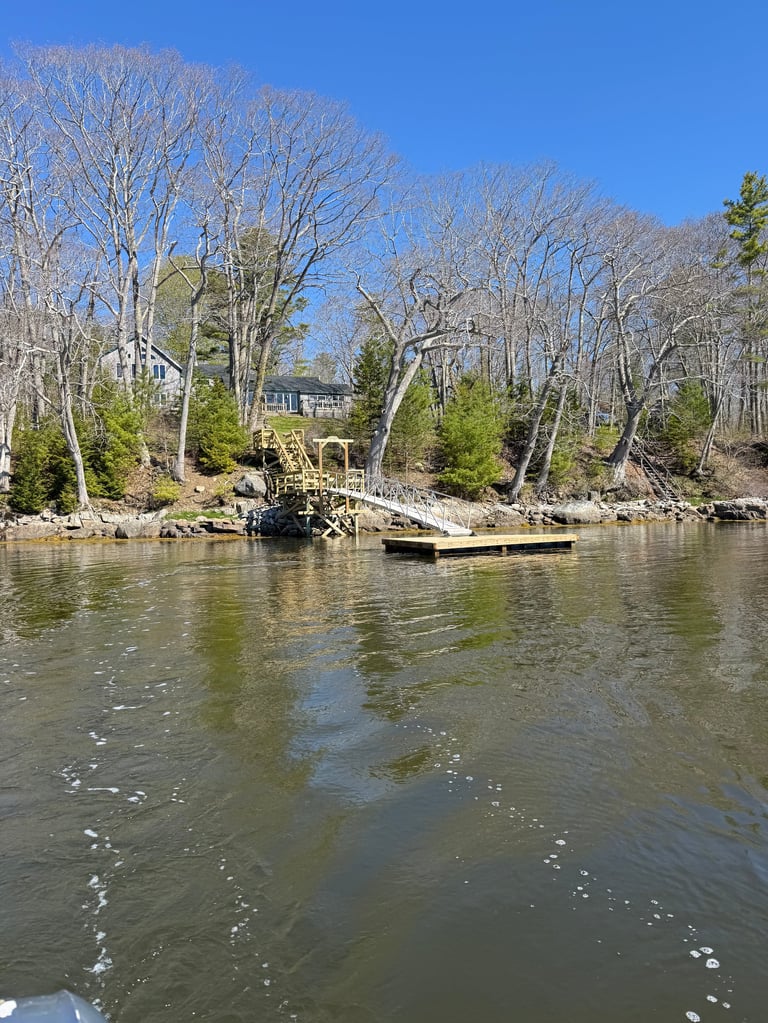Waterfront property with bare trees, dock, and yellow boathouse reflected in calm river water on clear sunny day